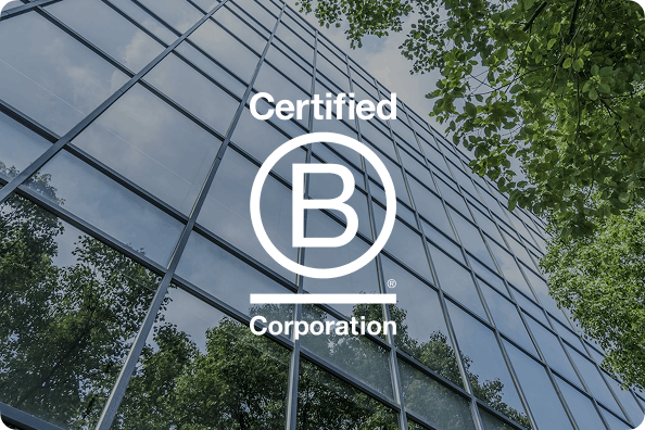 Modern glass building with geometric grid pattern, green trees visible at edges, white circular B Corp certification logo overlaid
