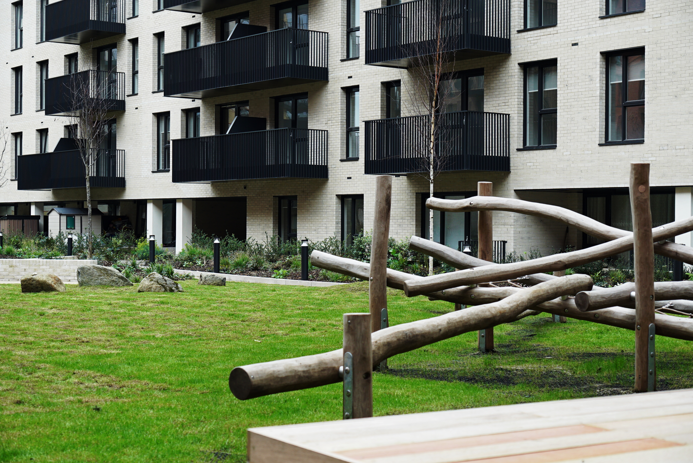 Wooden log climbing structure on a green lawn in a residential courtyard, with a modern apartment block featuring black balconies in the background.