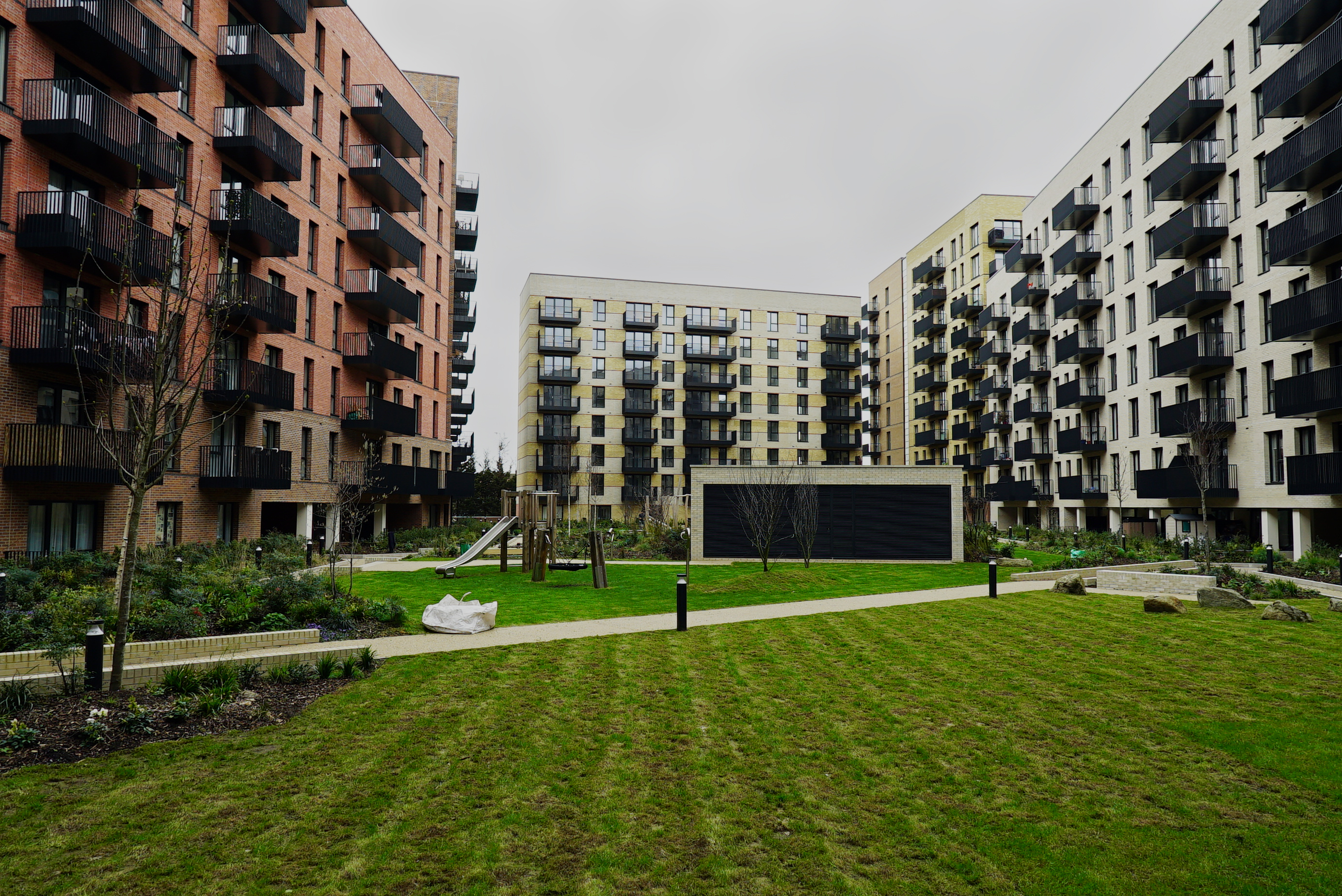 Modern residential apartment blocks with balconies surrounding a communal courtyard garden with green lawn and pathways, overcast sky.