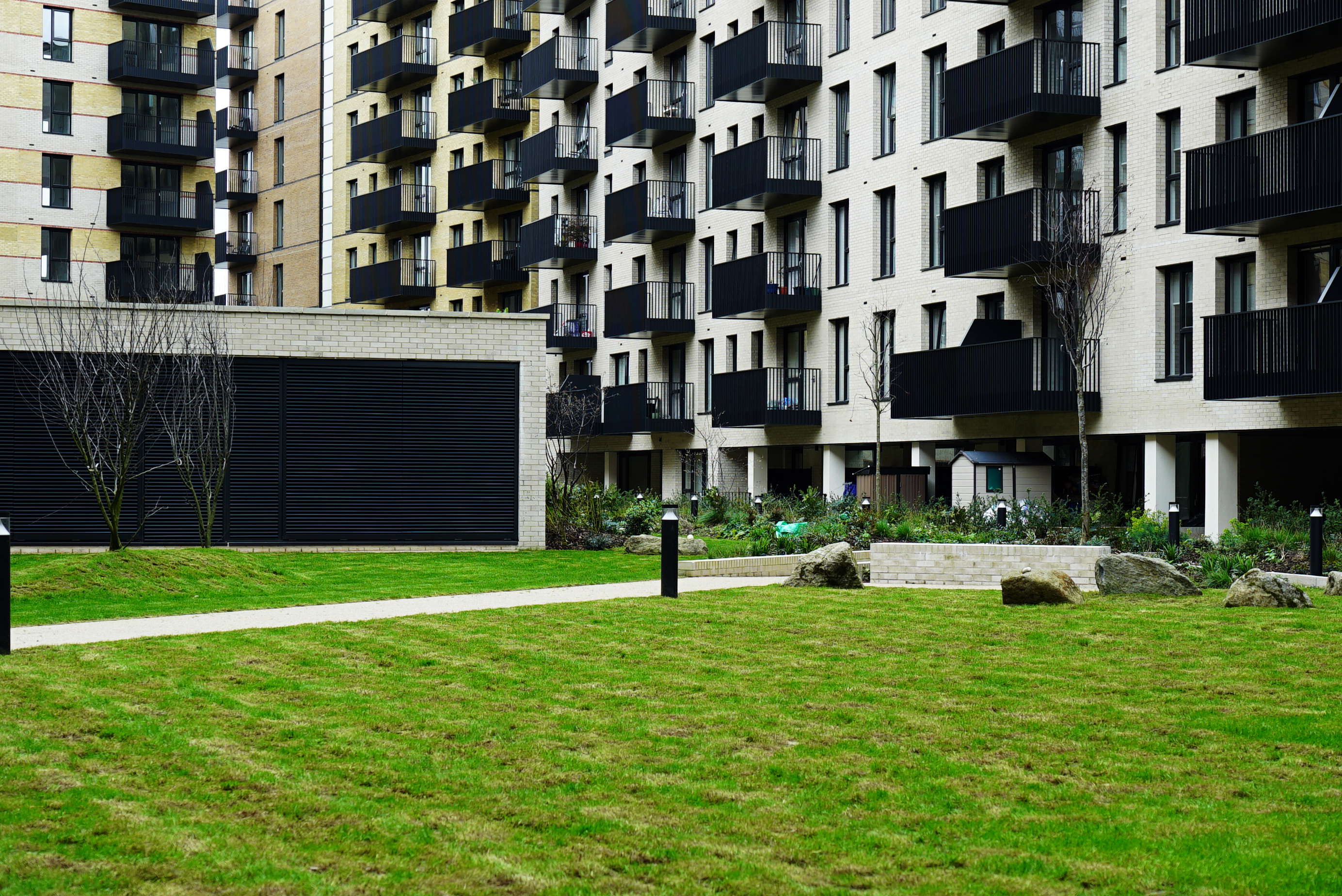 Modern residential apartment blocks with black balconies and cream facade, overlooking a communal green lawn with a footpath and rocks.