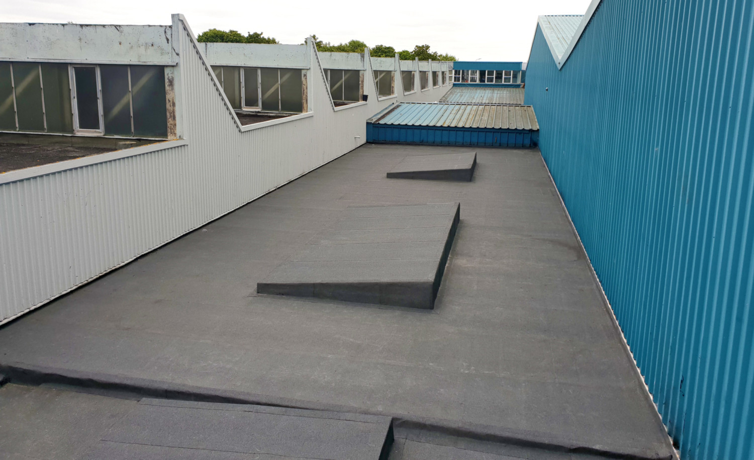 Industrial rooftop with grey concrete surface, teal corrugated metal wall on right, white building with windows on left, overcast sky.