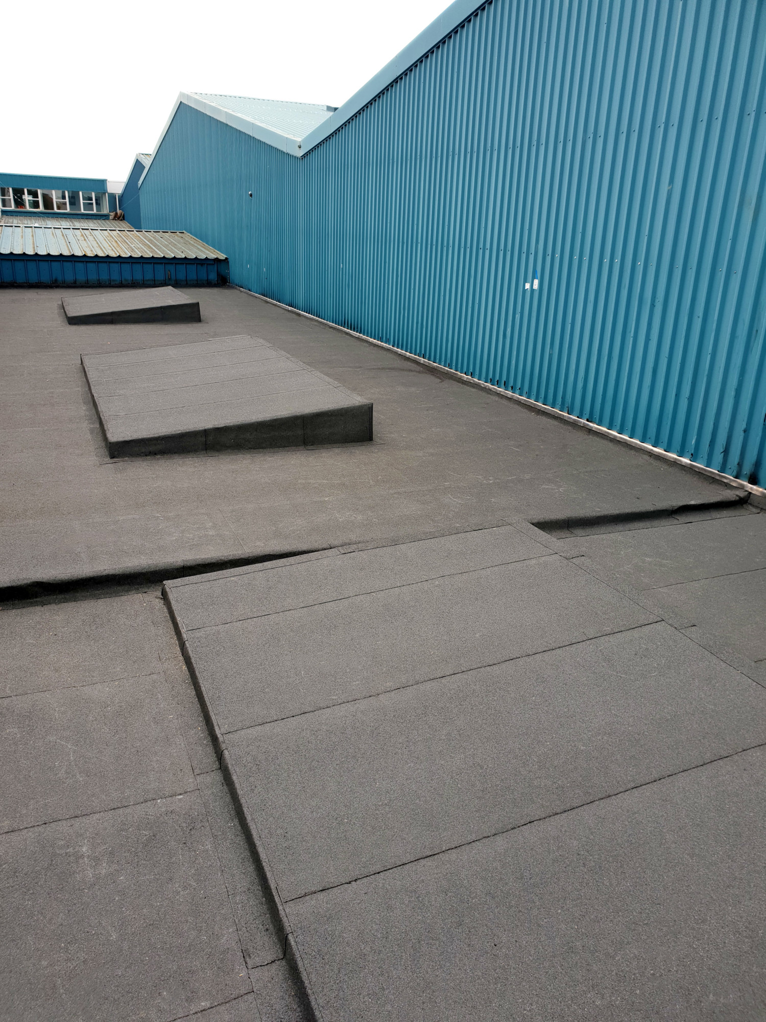Flat rooftop with grey concrete slabs and metal barriers alongside turquoise corrugated metal wall under overcast sky.