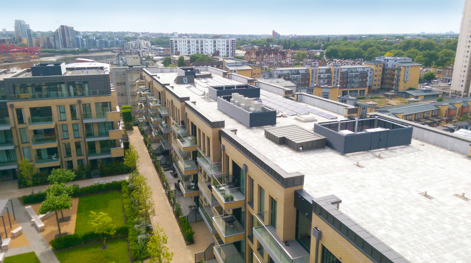 Aerial view of modern residential development with beige buildings, white rooftops, green spaces, and urban skyline in background.