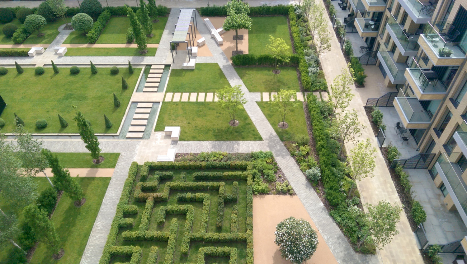Aerial view of landscaped courtyard with geometric hedge maze, green lawns, concrete pathways, and modern residential buildings.