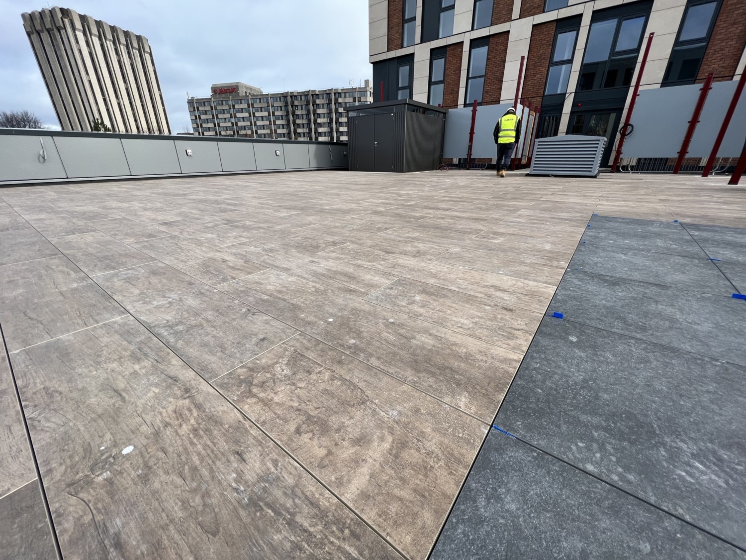Concrete rooftop with geometric paving pattern, modern brick buildings in background, worker in high-vis jacket visible.