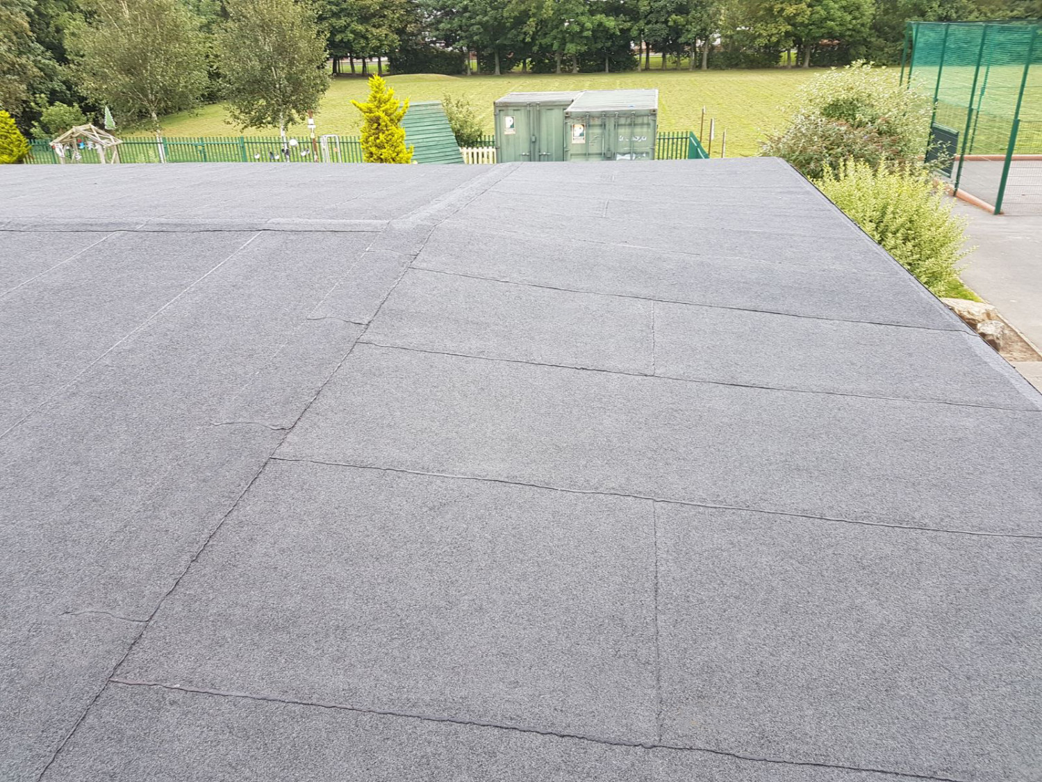Grey flat roof surface with visible panel joints, surrounded by green trees, grass field, and sports court fencing in background.