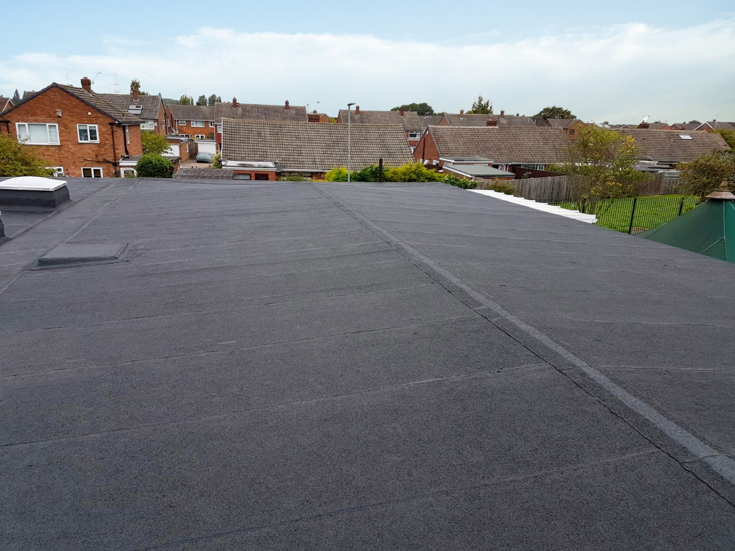 Dark grey flat roof with visible seams and drainage channels, overlooking residential area with red brick houses and tiled roofs.