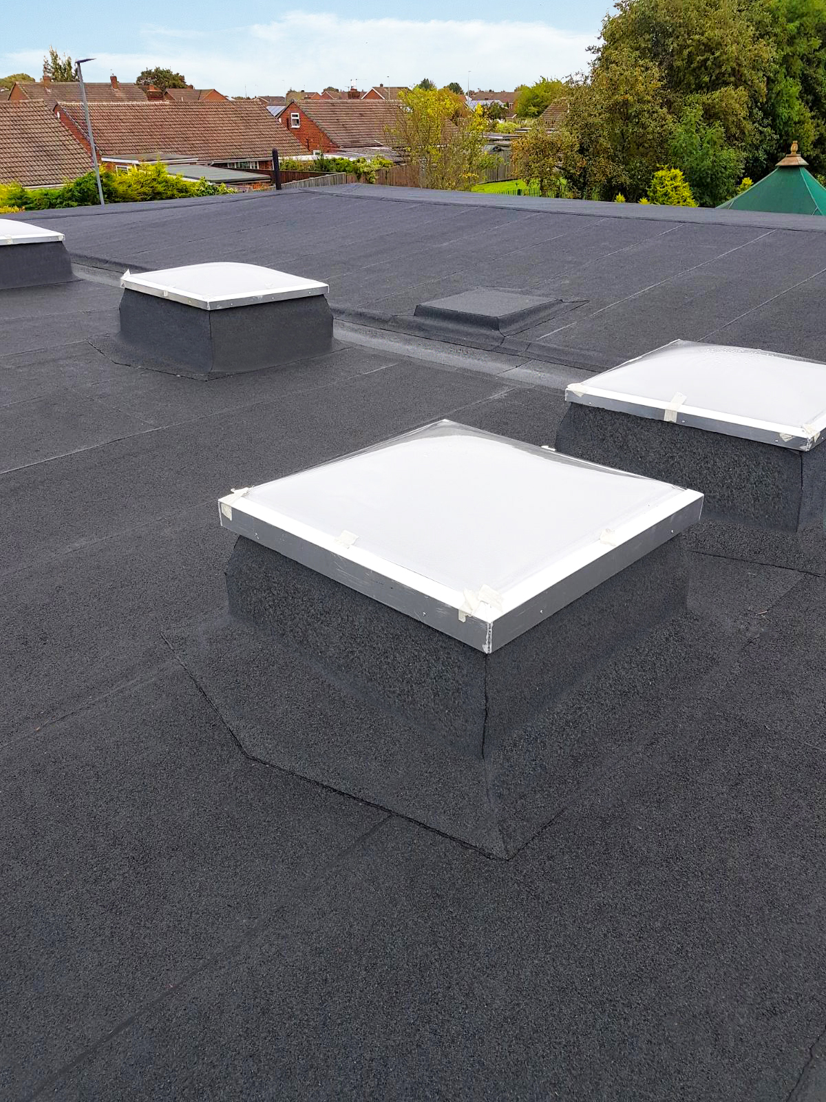 Flat roof with four white square rooflights on dark membrane surface, residential area with houses and trees visible beyond.