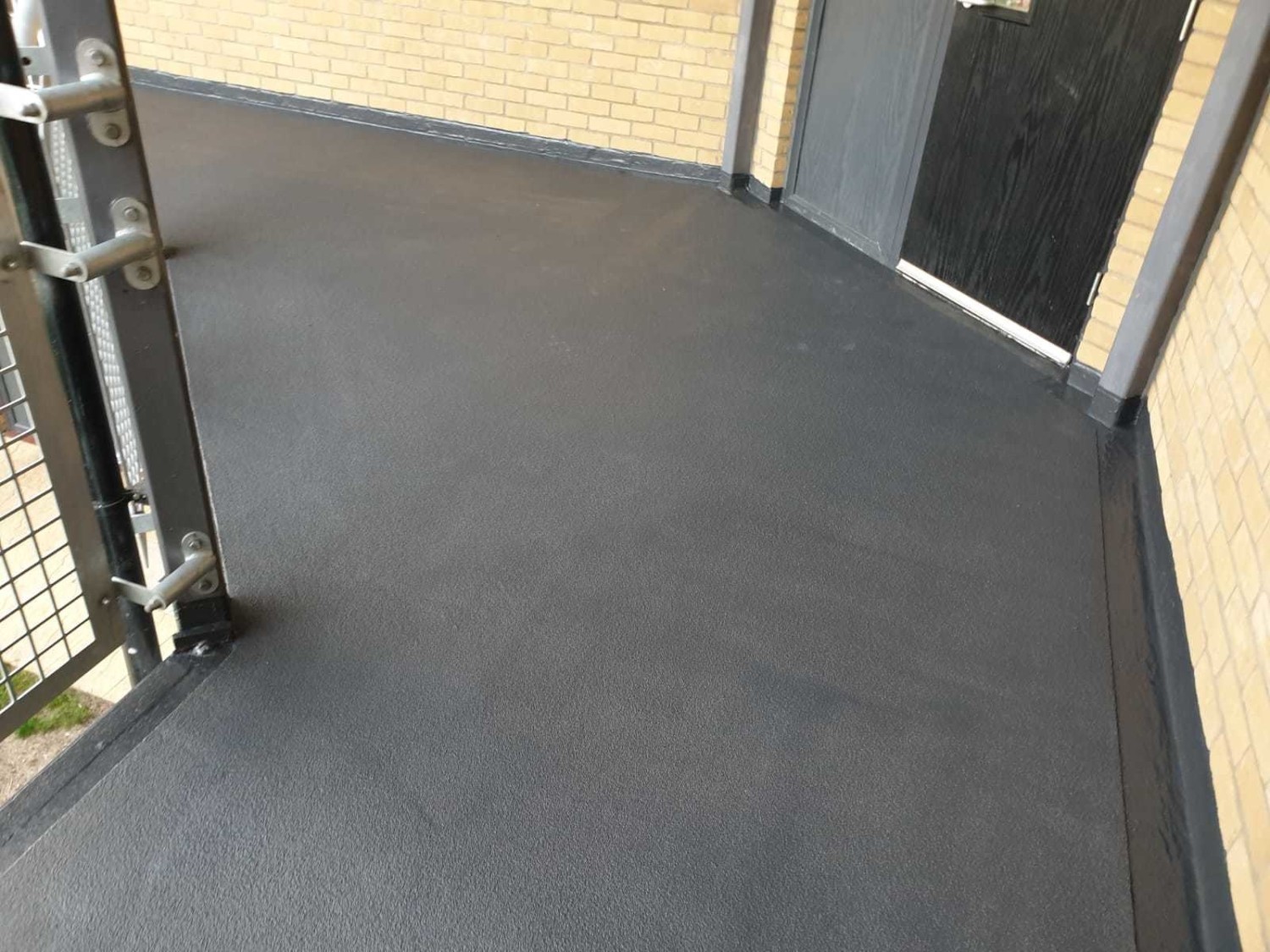 Dark grey waterproof membrane on flat roof with metal railings and cream-coloured building walls visible at edges.