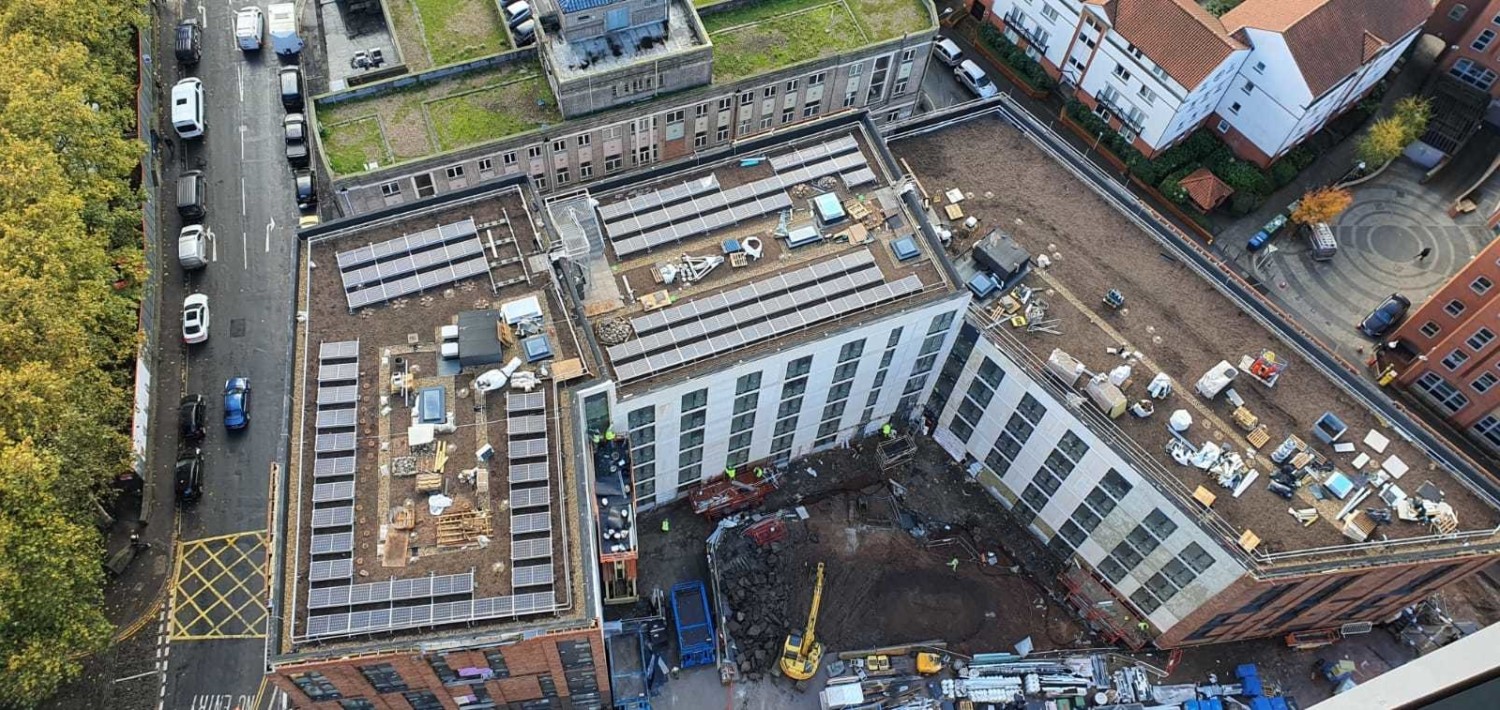 Aerial view of construction site showing concrete foundation work, white multi-storey building frame, brown earth, green trees, and surrounding residential houses with red roofs.