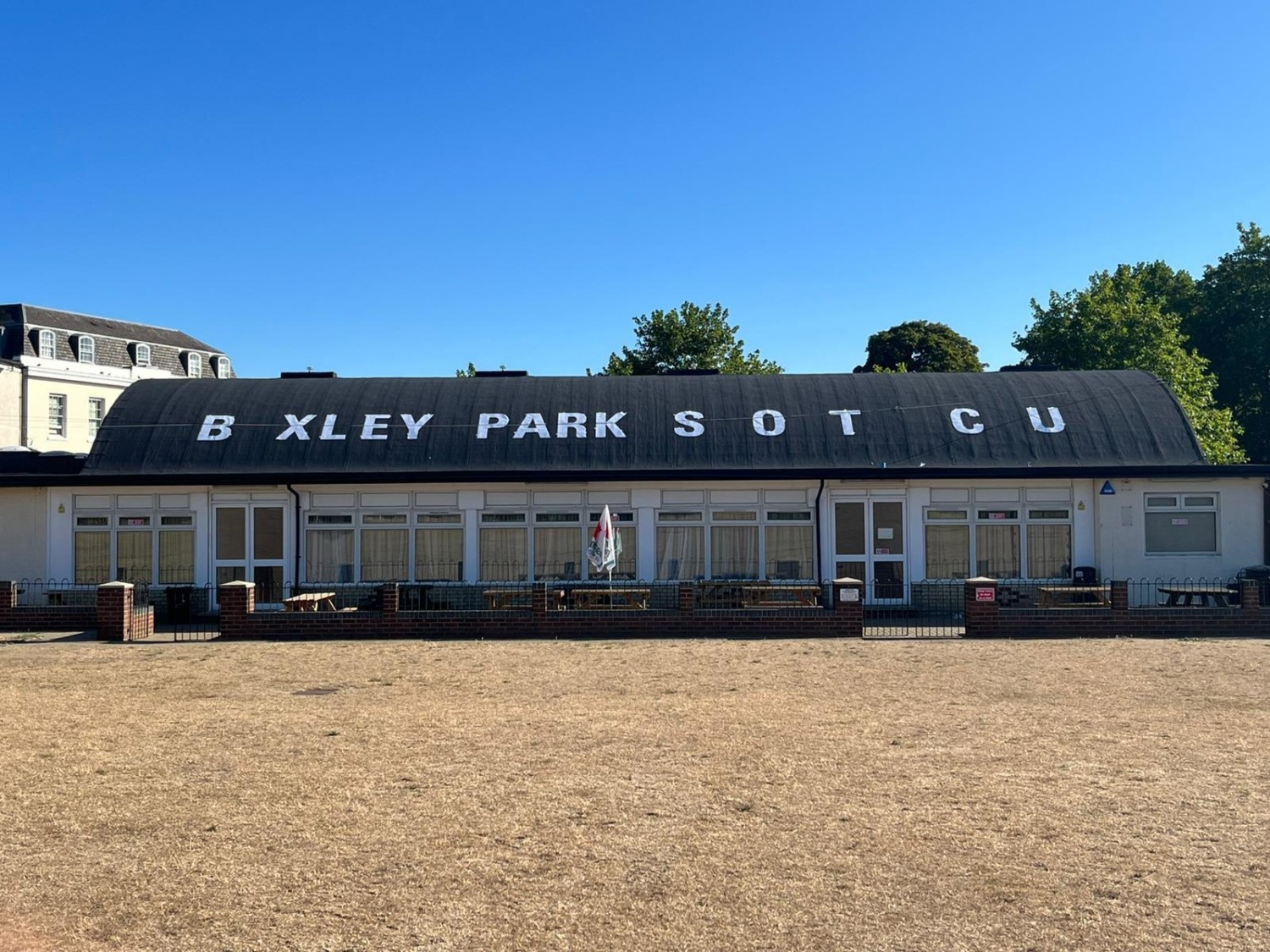 Single-storey white pavilion building with dark roof displaying "BEXLEY PARK S O T C U" in large white letters against blue sky.