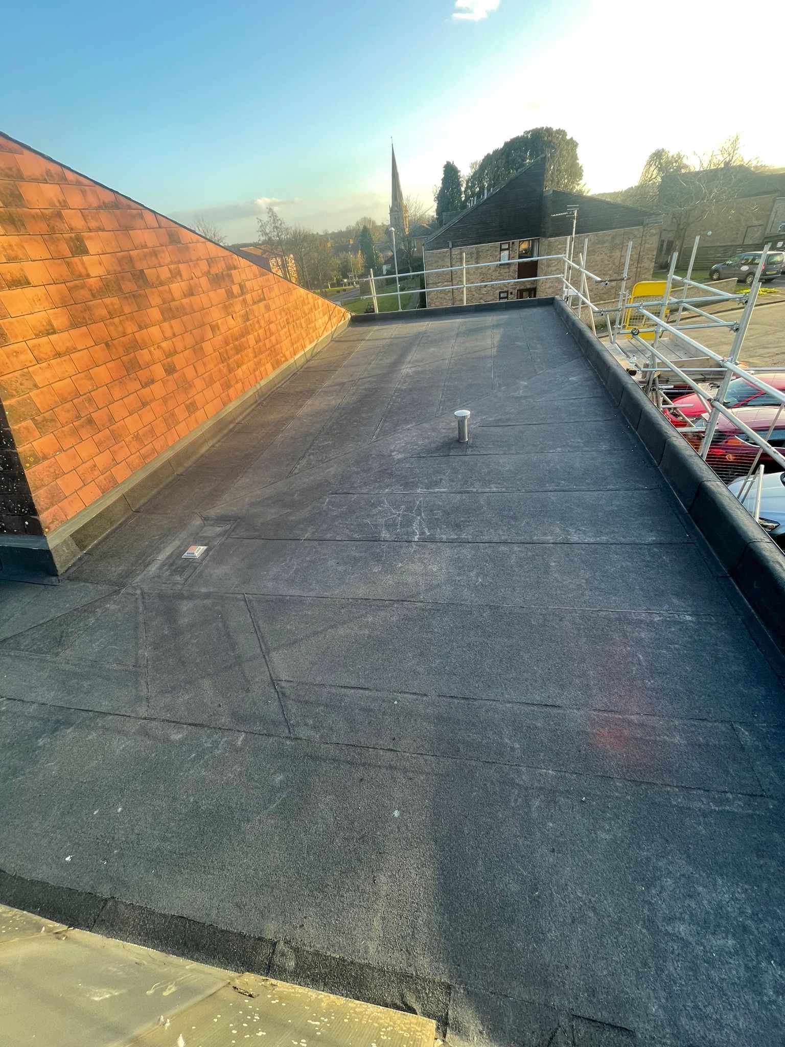 Flat roof with dark membrane, orange clay tile wall on left, white scaffolding, church spire and trees visible in background.