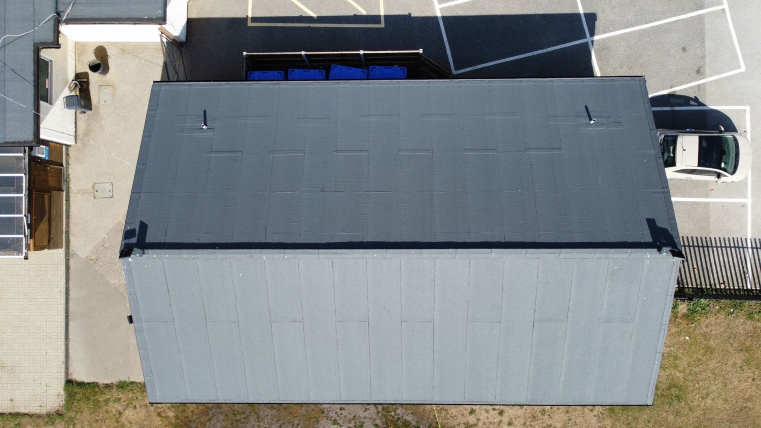 Aerial view of rectangular grey roof with white building sections, blue pool, and concrete surfaces surrounding property.