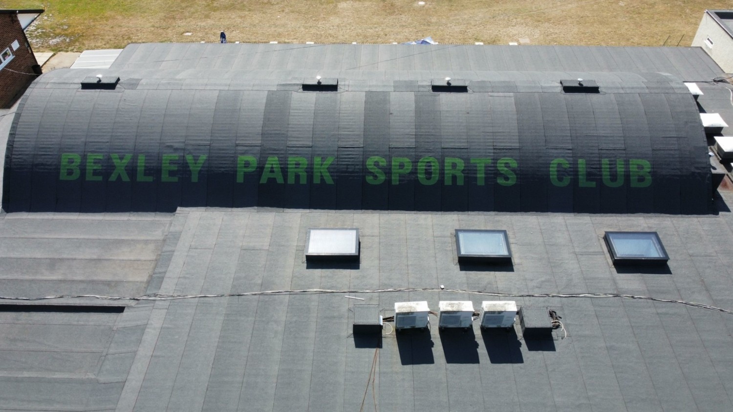 Aerial view of large green hangar building with "BEACON PARK SPORTS CLUB" text on curved roof, surrounded by concrete apron.