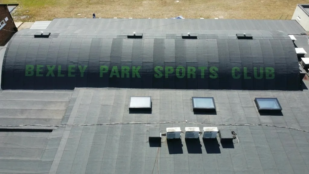 Aerial view of large curved green building with "BEXLEY PARK SPORTS CLUB" text on roof, surrounded by grey concrete areas.