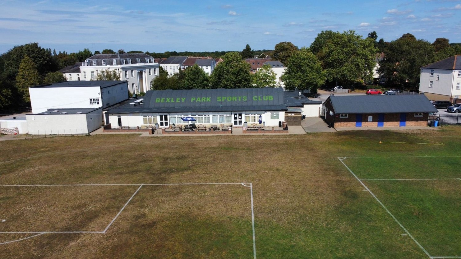 Aerial view of school buildings with sports fields. Brown and green pitches in foreground, grey and white buildings behind, surrounded by trees and houses under blue sky.