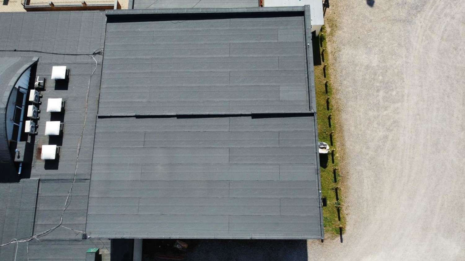 Aerial view of grey industrial building with flat roof next to beige concrete surface, separated by narrow strip of grass.