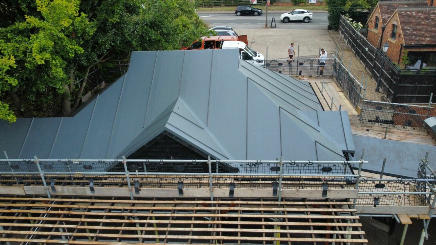 Aerial view of building construction with grey metal roofing, wooden framework, scaffolding, and workers on site surrounded by trees.