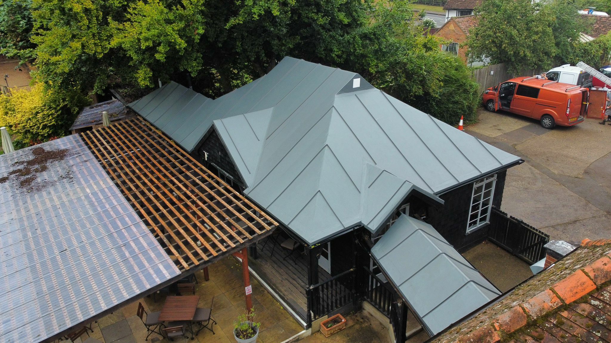 Aerial view of house with grey metal roof sections and exposed wooden roof framing during construction work.