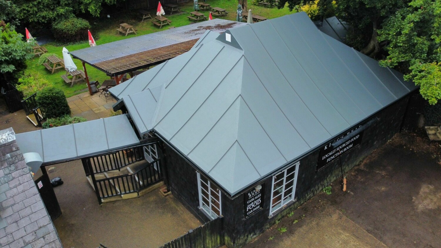 Aerial view of dark wooden building with grey metal roof, covered walkway, and wooden decking surrounded by green trees.