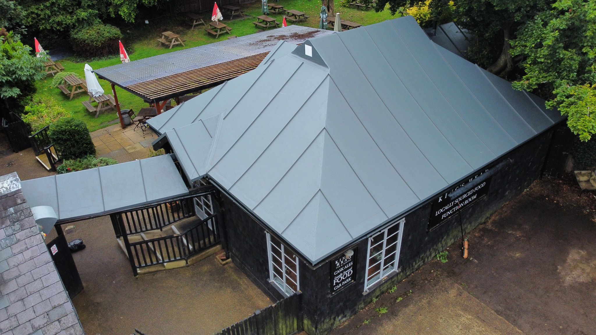 Aerial view of dark wooden building with grey metal roof, wooden deck extension, surrounded by green trees and gravel paths.