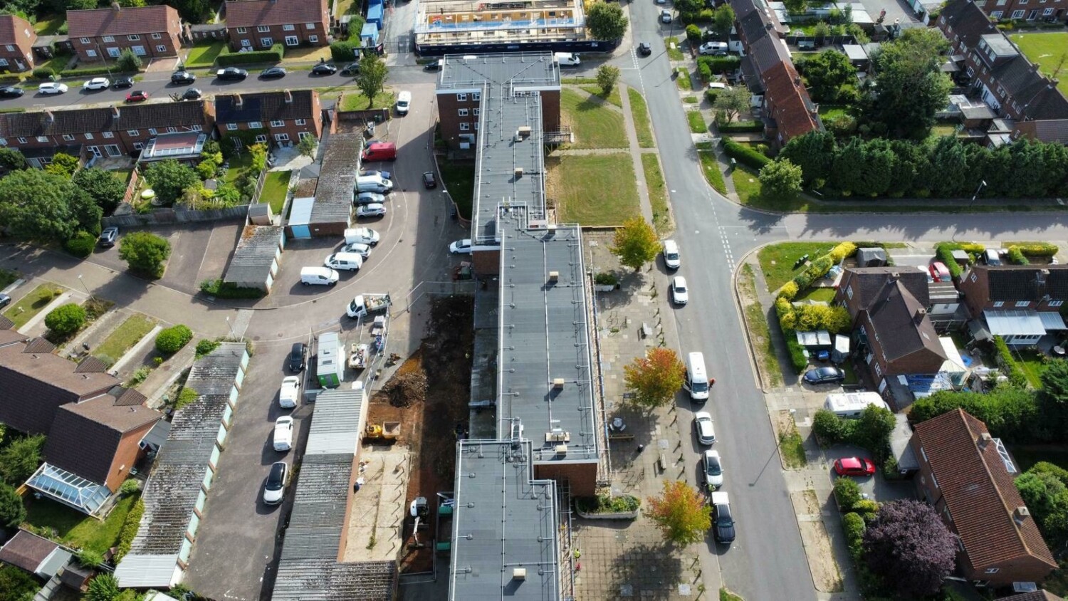 Aerial view of residential street with grey rooftops, green gardens, parked cars, and tree-lined roads forming grid pattern.
