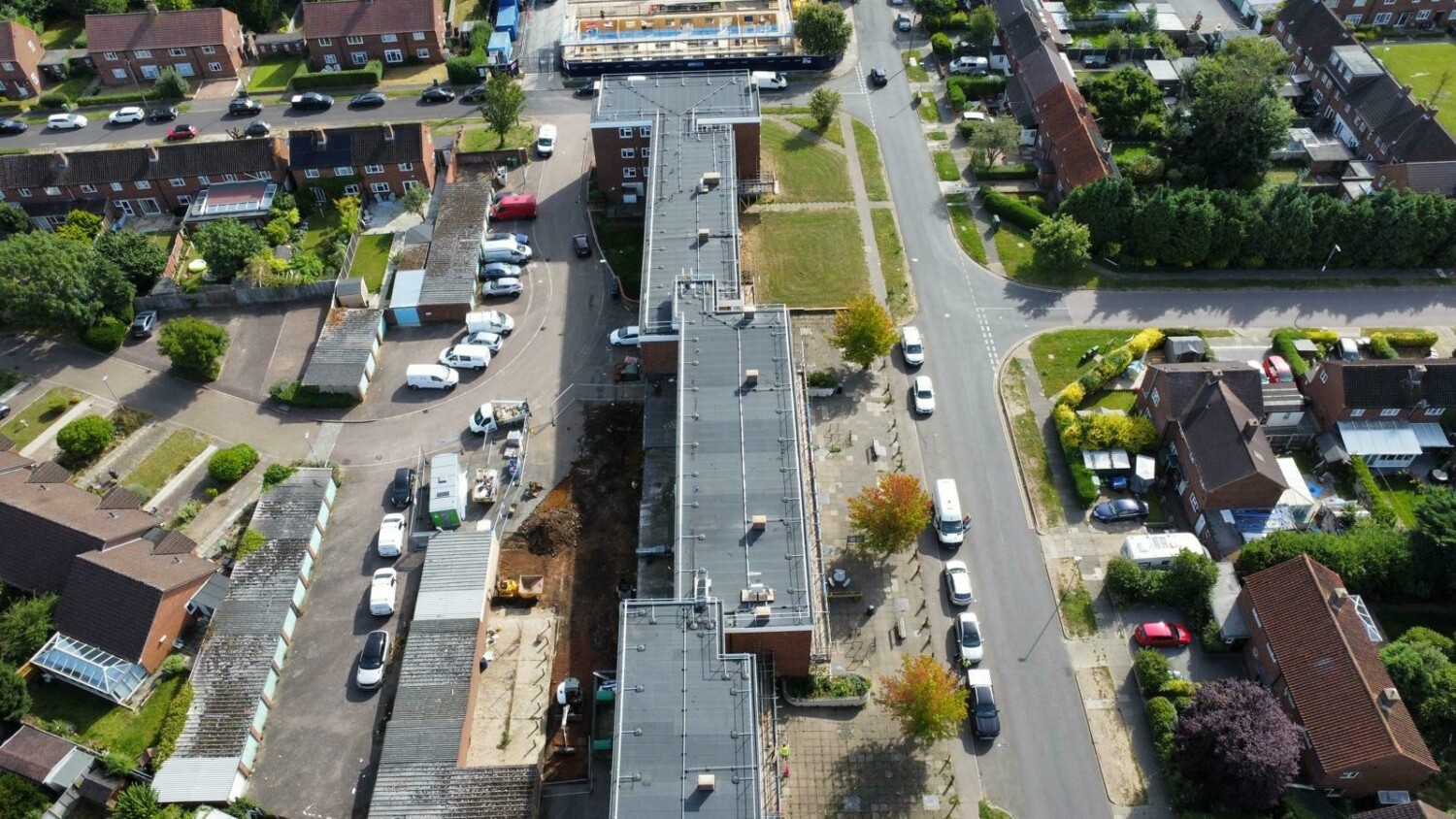 Aerial view of residential street with grey road, parked cars, houses with varied roof colours, green trees, and grassy areas.