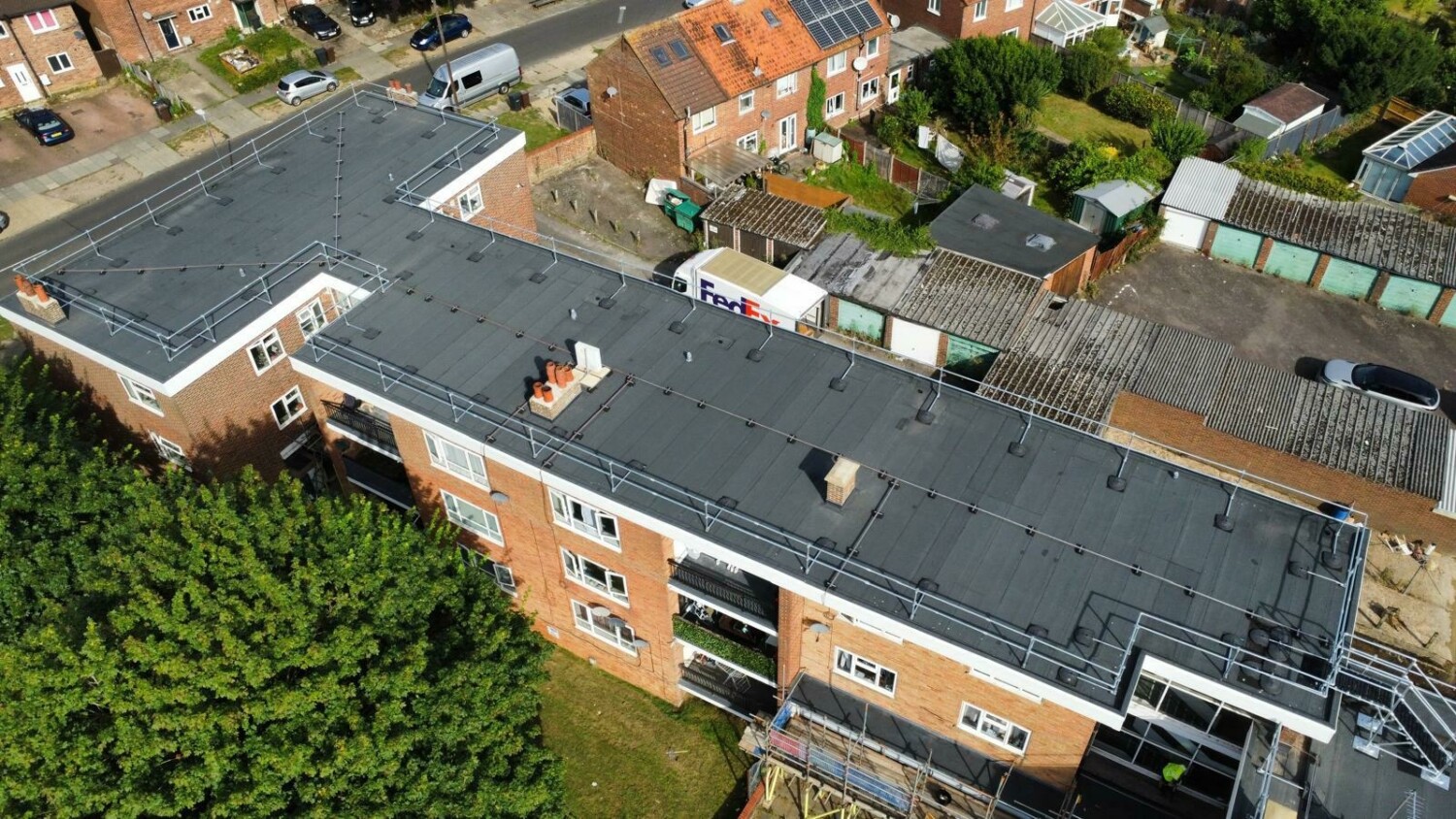 Aerial view of brick residential building with dark grey flat roof surrounded by houses, gardens, and parked cars in suburban area.