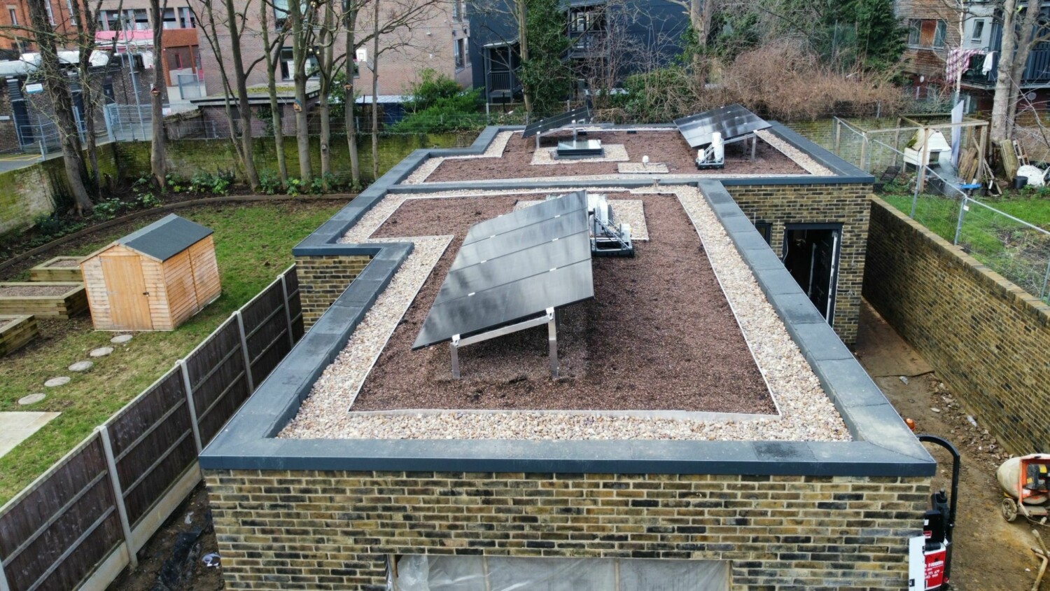 Aerial view of partially constructed flat-roofed building with brick walls, grey roofing sections, and brown material covering areas.