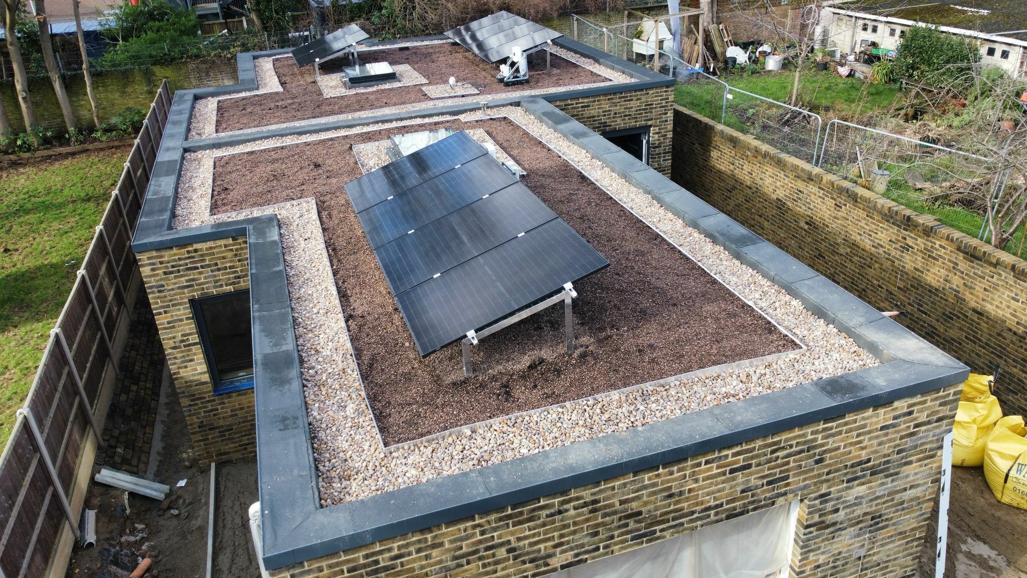 Aerial view of flat roof with dark solar panels installed on brown gravel surface, surrounded by brick walls and residential area.