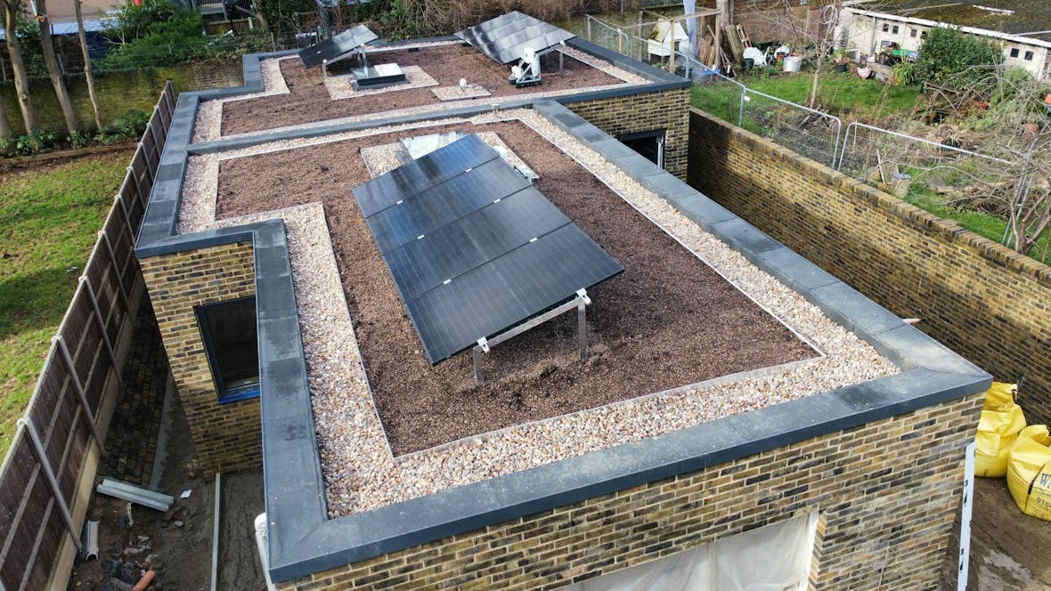 Aerial view of flat roof with dark solar panels installed on brown surface, brick walls visible, surrounded by residential area.