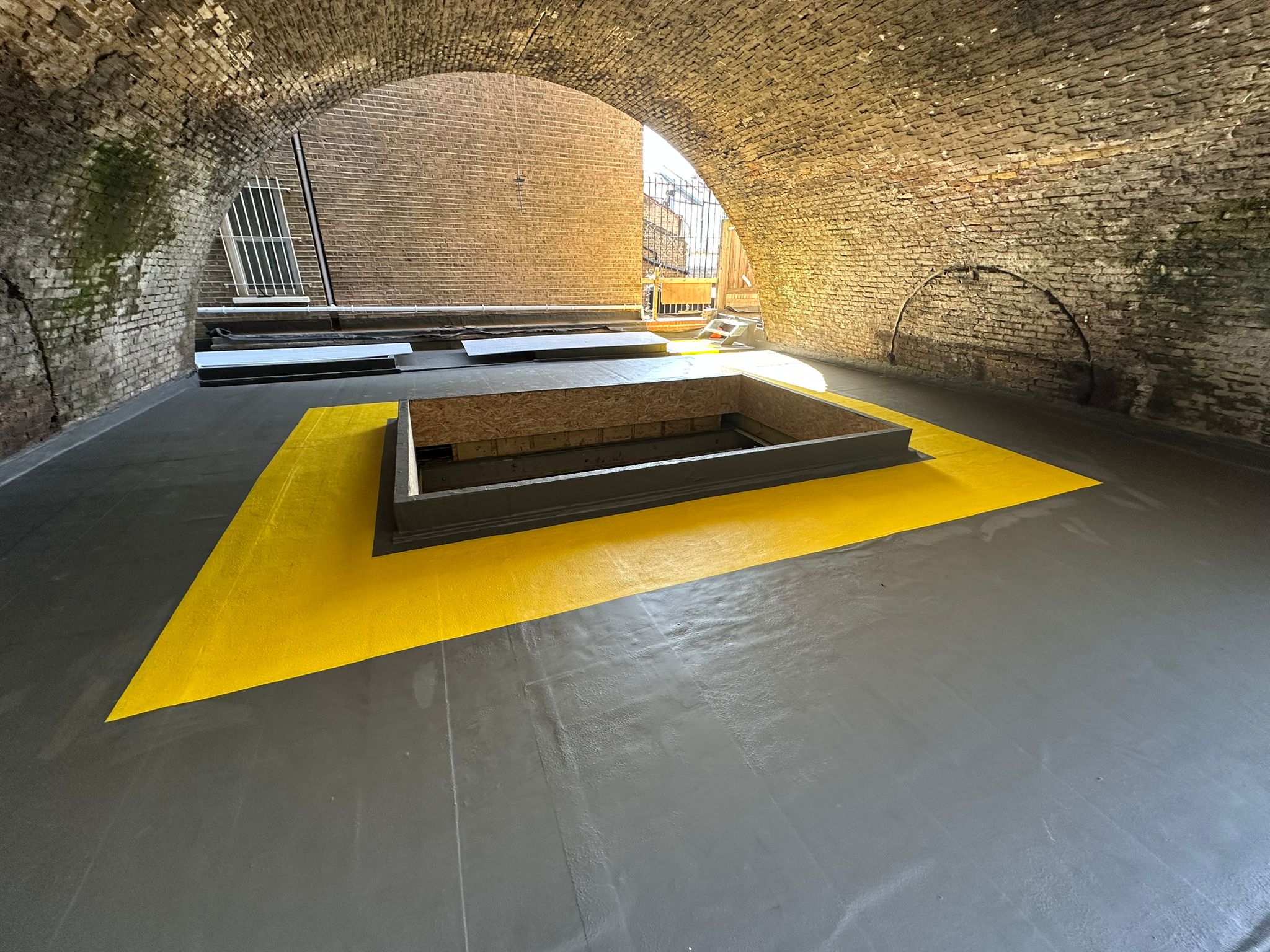 Underground brick-vaulted space with grey floor, bright yellow triangular surface, dark rectangular platform, and metal railings.