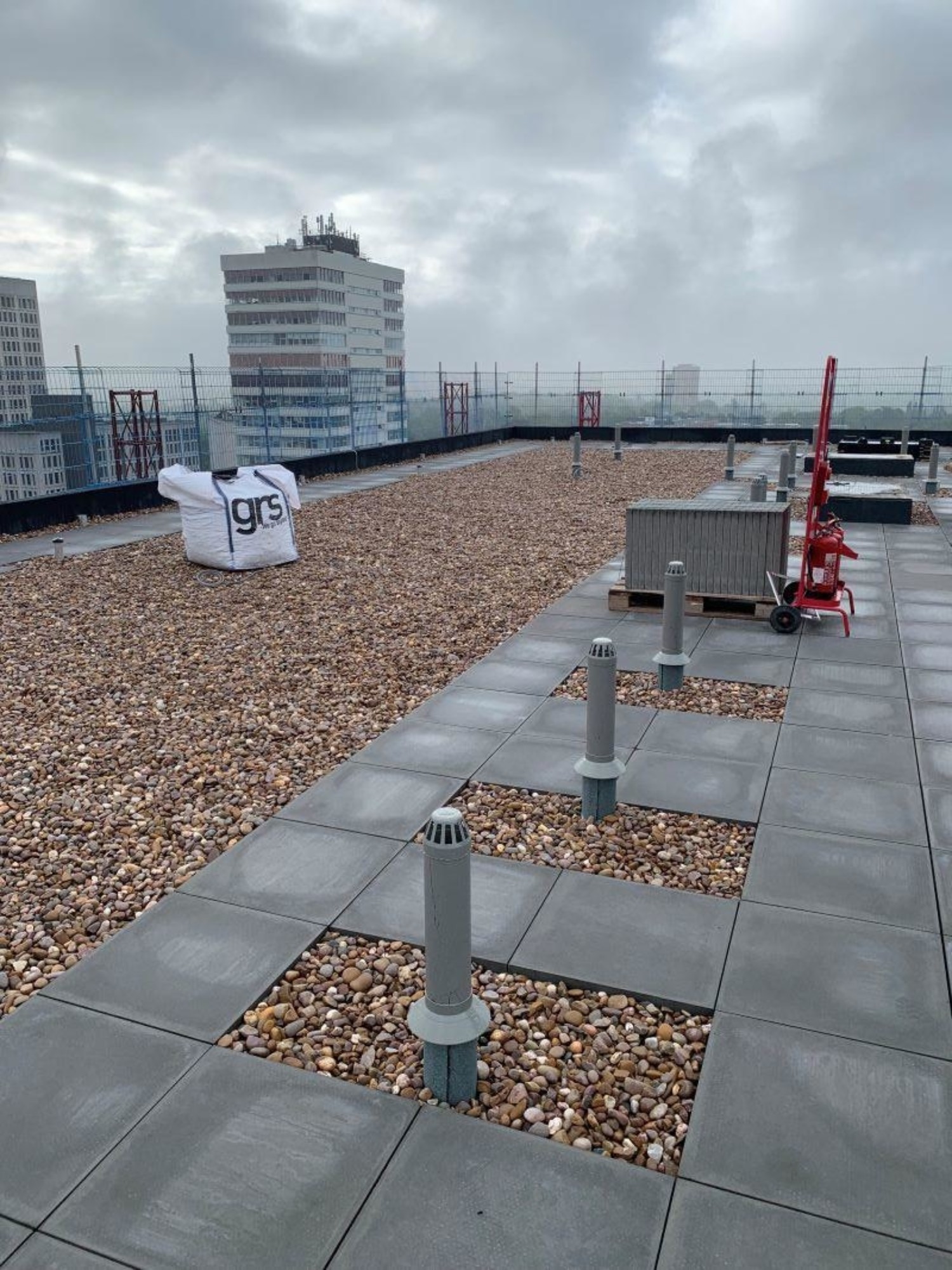 Rooftop with grey concrete slabs and brown gravel sections. White inflatable cube with "gr3" text. Red crane and buildings visible in background under cloudy sky.