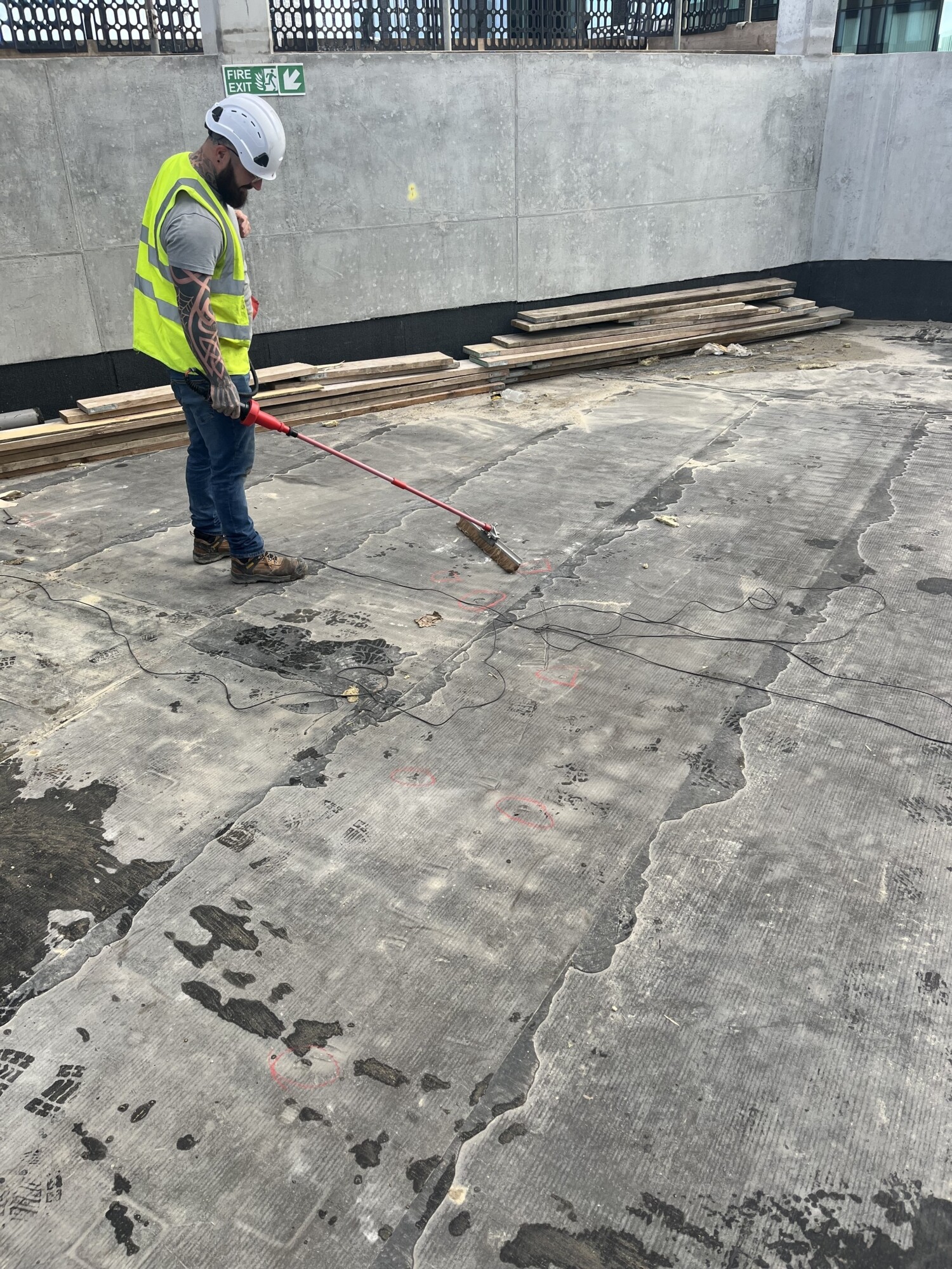 Construction worker in high-vis jacket and hard hat using long-handled tool on damaged concrete floor with wooden planks nearby.