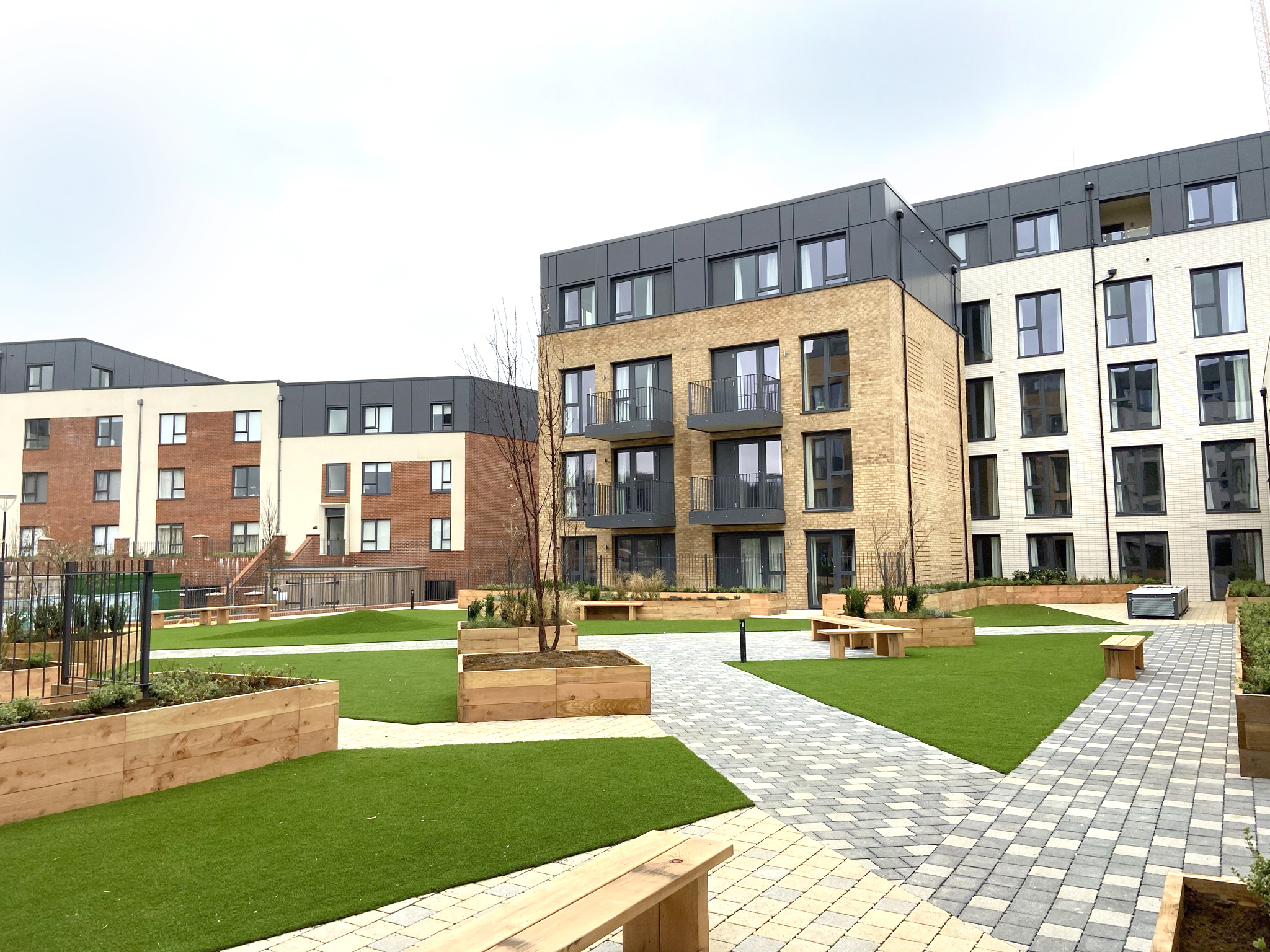 Modern residential development with multi-storey brick and grey buildings surrounding landscaped courtyard with green lawns, paved paths, and wooden benches.