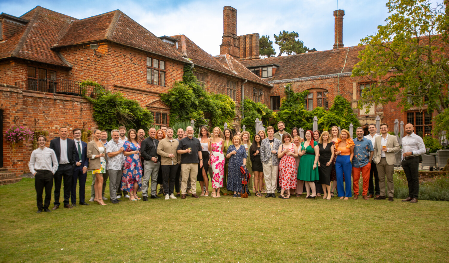 Large group of people in formal attire standing on green lawn before red brick Tudor-style building with multiple chimneys.