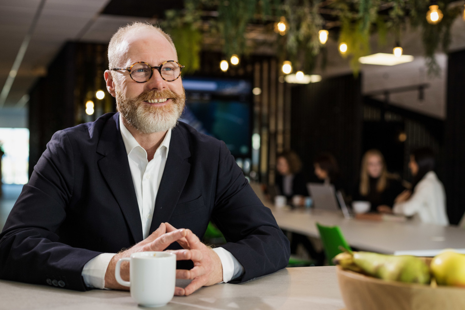 Smiling bearded man with glasses in dark suit jacket sits at table with white mug, modern office with warm lighting behind.