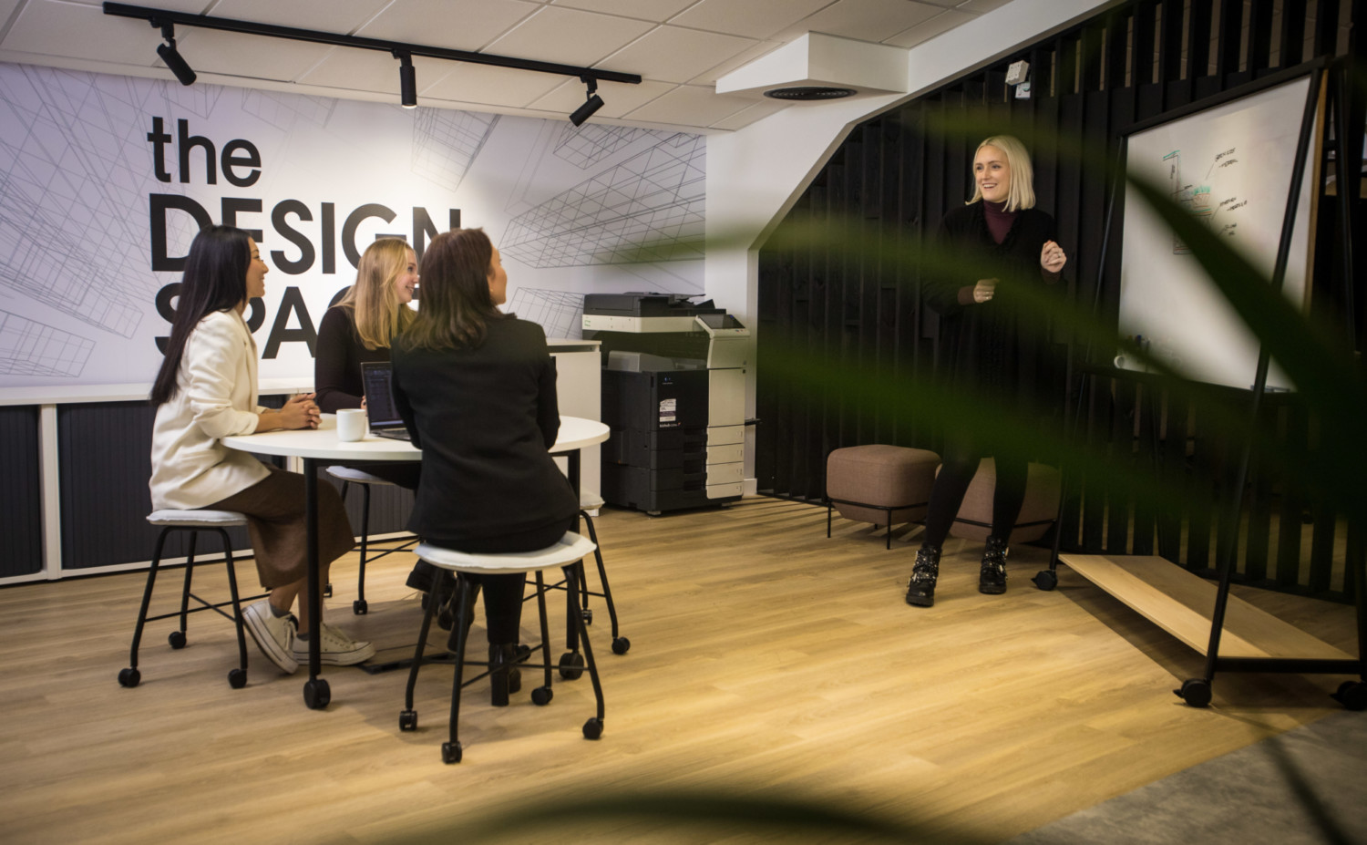 Woman presenting to two seated people in modern office space with "the DESIGN" wall text, projector screen, and wooden flooring.