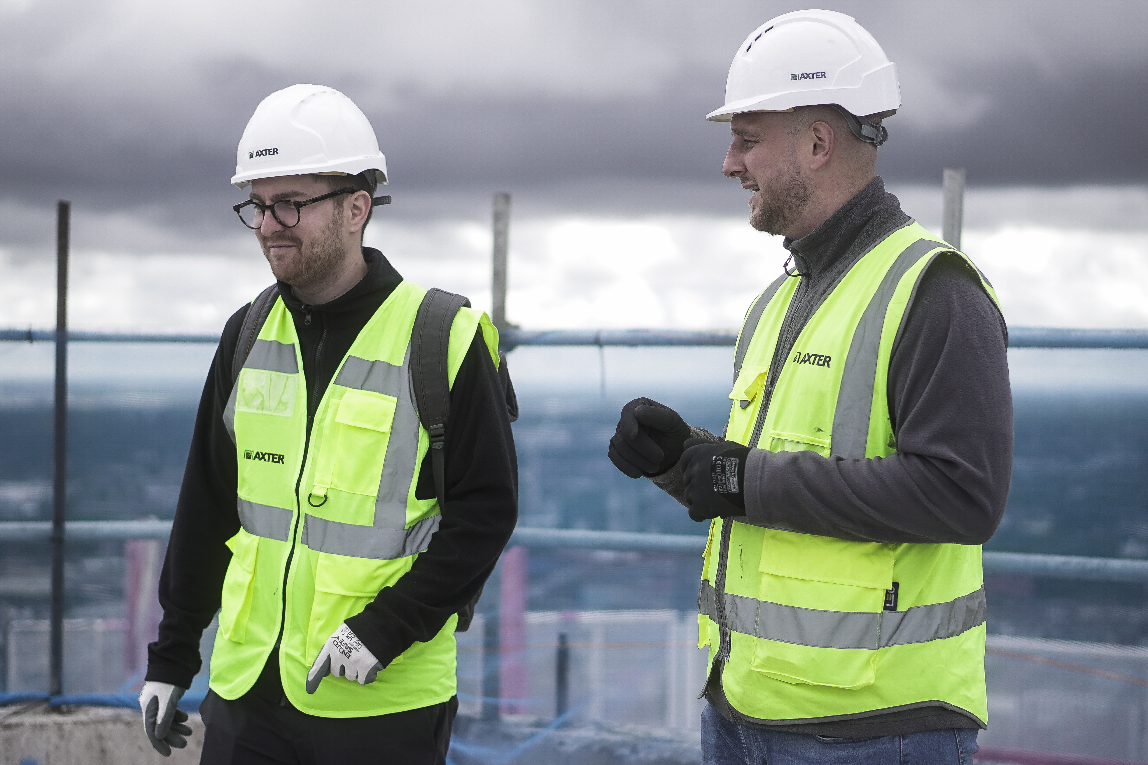 Two construction workers in high-vis yellow vests and white hard hats conversing on building site with grey cloudy sky behind.