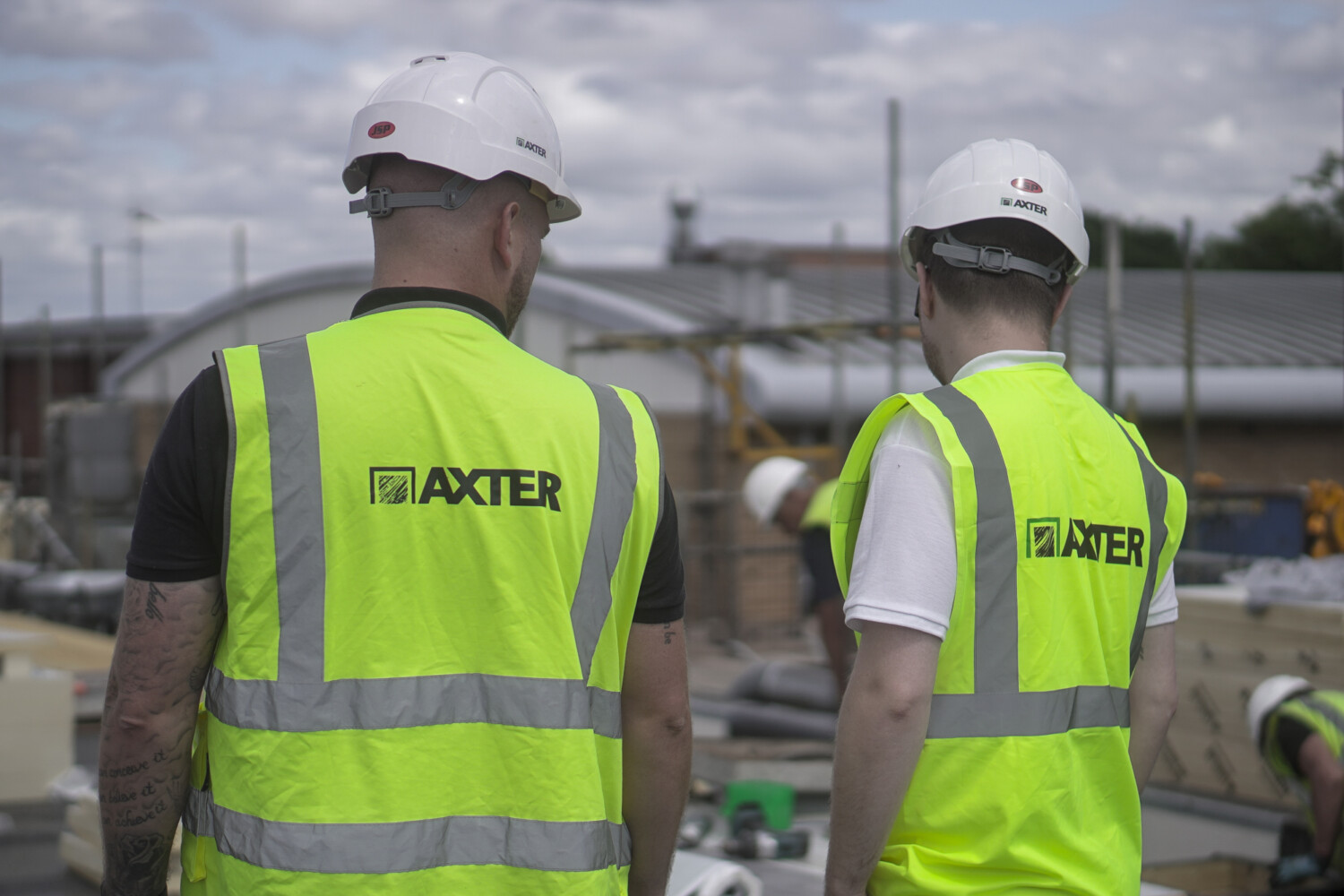 Two construction workers in white hard hats and bright yellow high-vis vests with "AXTER" branding, viewed from behind at building site.