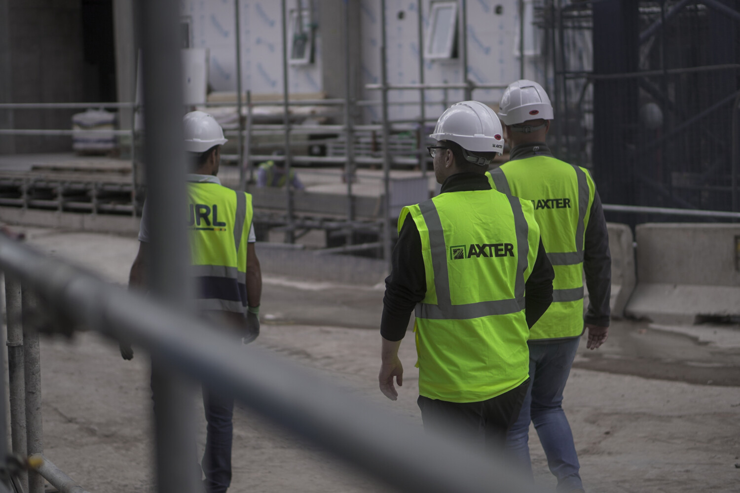 Three construction workers in high-vis yellow jackets and white hard hats walking through concrete building site with scaffolding.
