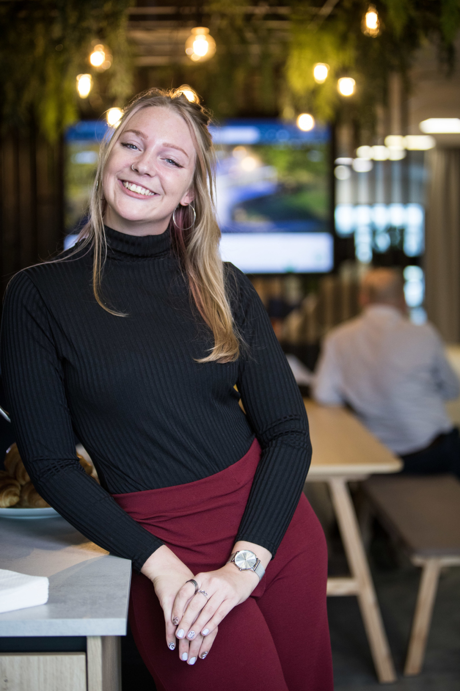 Young blonde woman in black turtleneck and burgundy skirt smiling in restaurant with warm lighting and blurred diners behind.