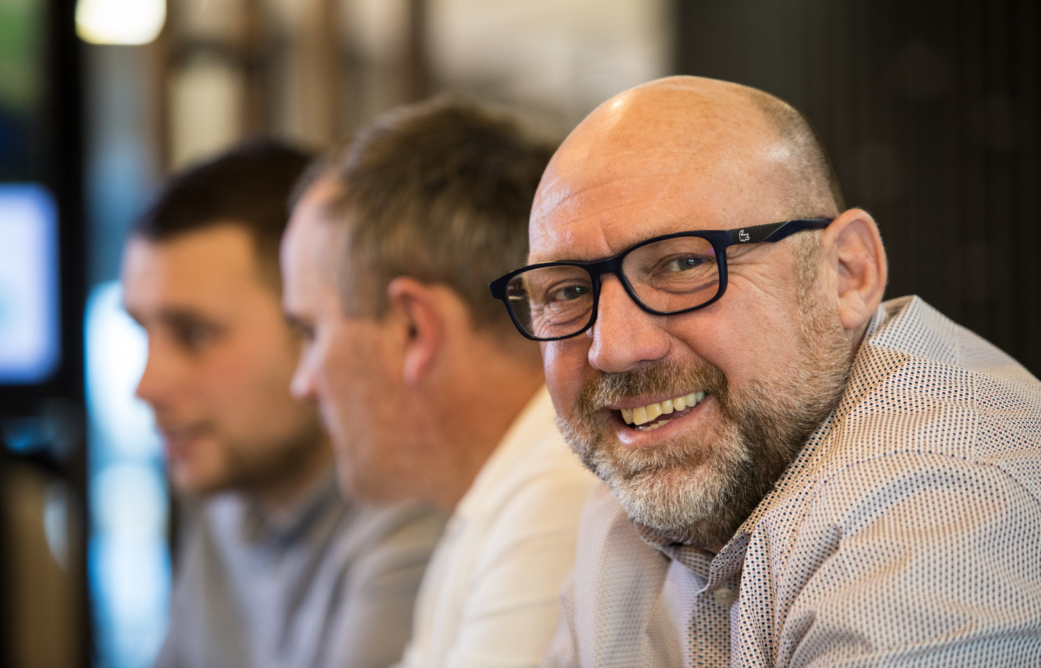 Bald man with glasses and beard wearing striped shirt, smiling at camera with two other men blurred in background indoors.