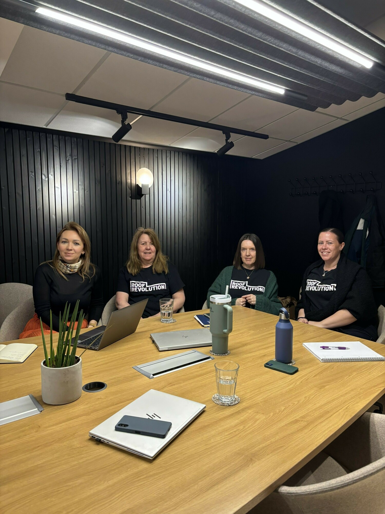 Four women seated at wooden conference table in modern office. Three wear matching dark t-shirts. Documents, laptops, and water glasses on table.