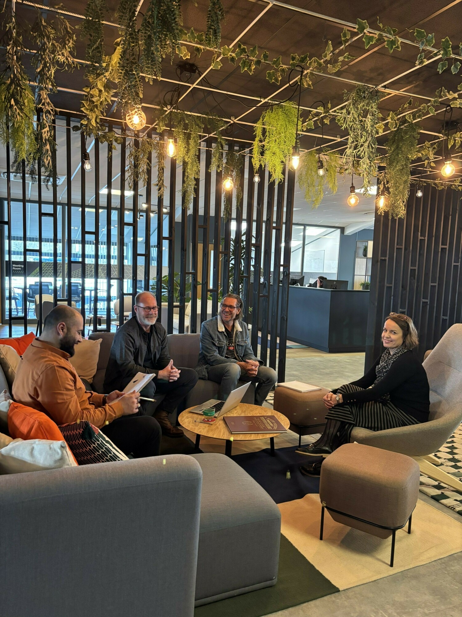Four people sitting in modern office lounge with grey furniture, hanging plants, Edison bulbs, and vertical wood slat dividers.