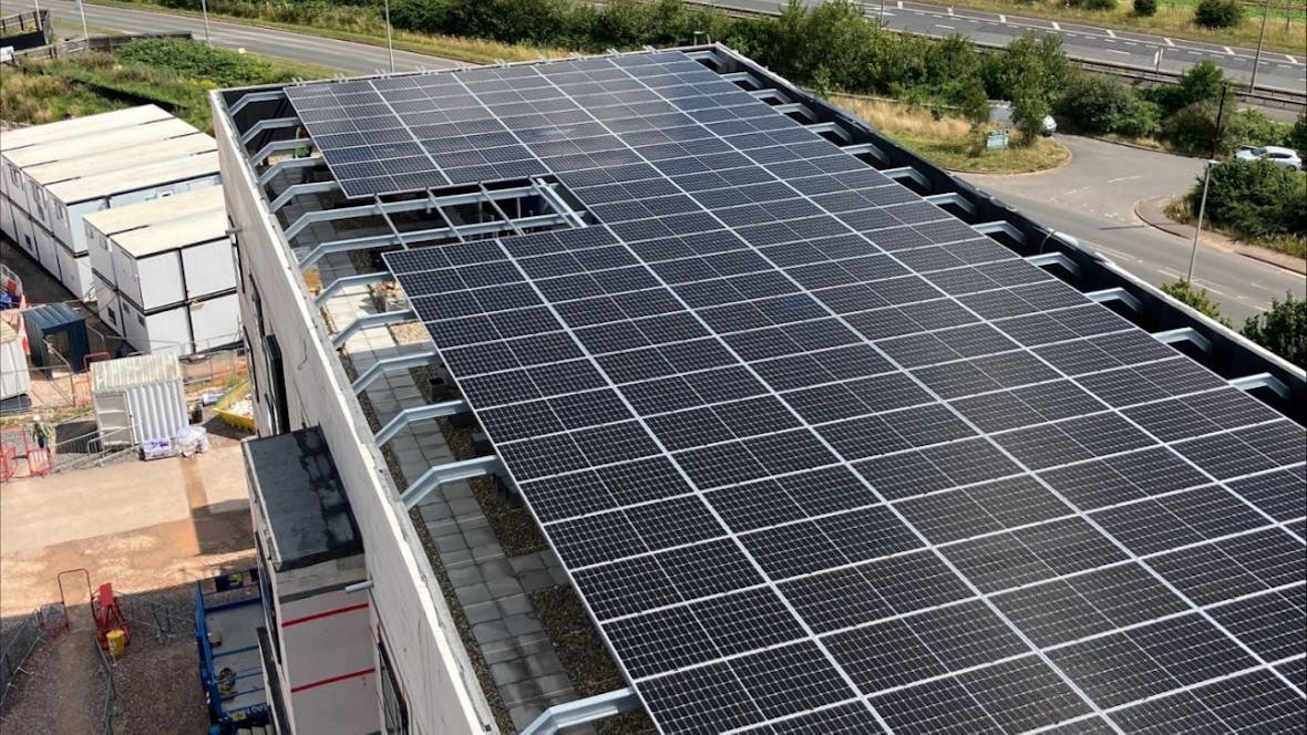 Large solar panel array covering industrial building rooftop, with surrounding car park and green spaces visible from aerial view.