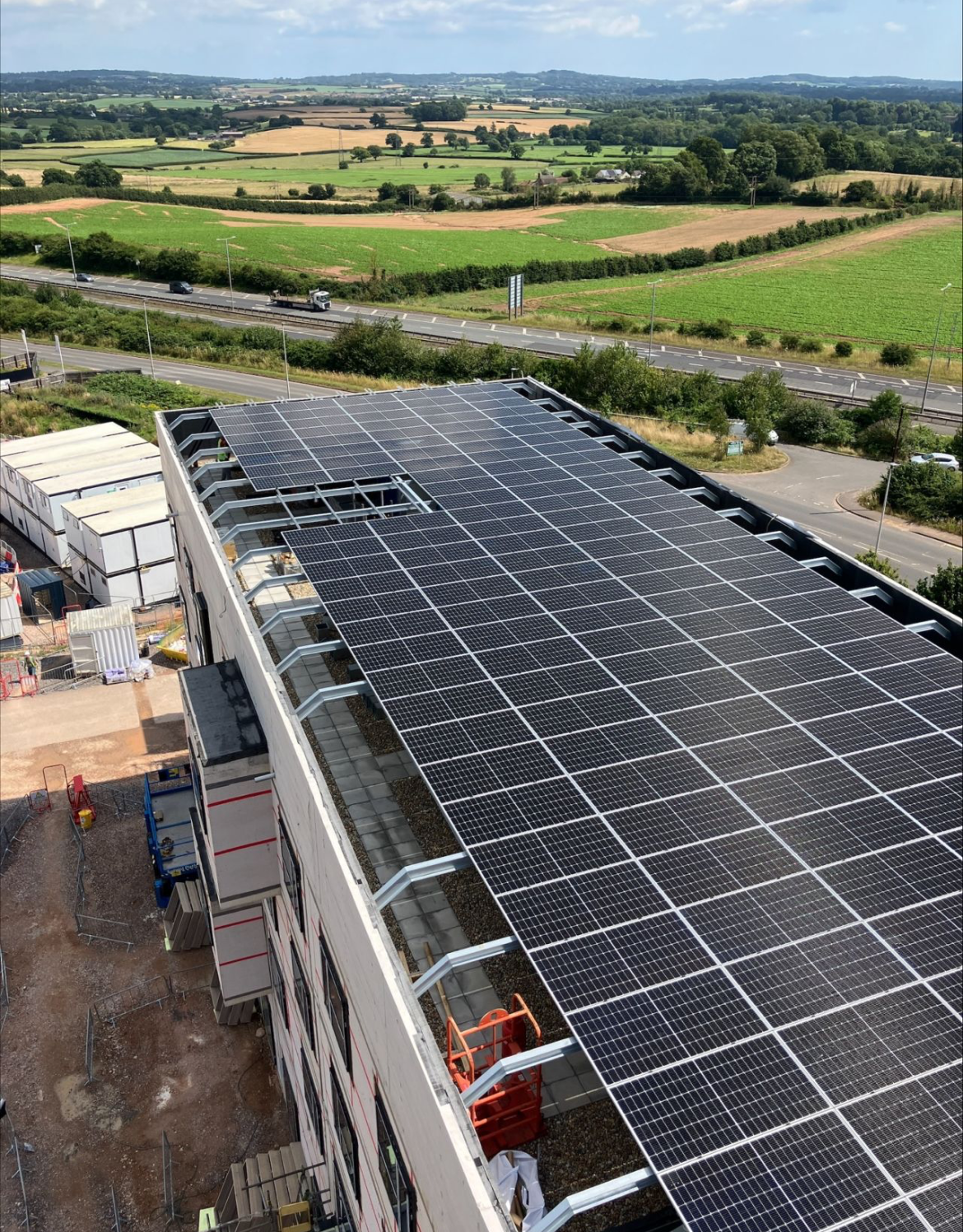 Large solar panel array on industrial building roof with countryside fields, road and hills in background.