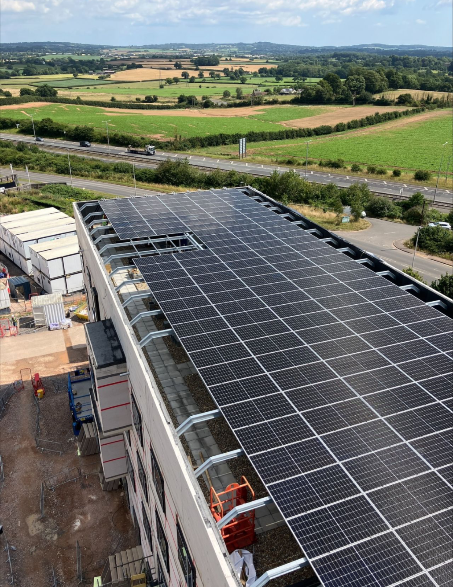 Aerial view of large industrial building with solar panels covering entire roof, surrounded by green fields and countryside.