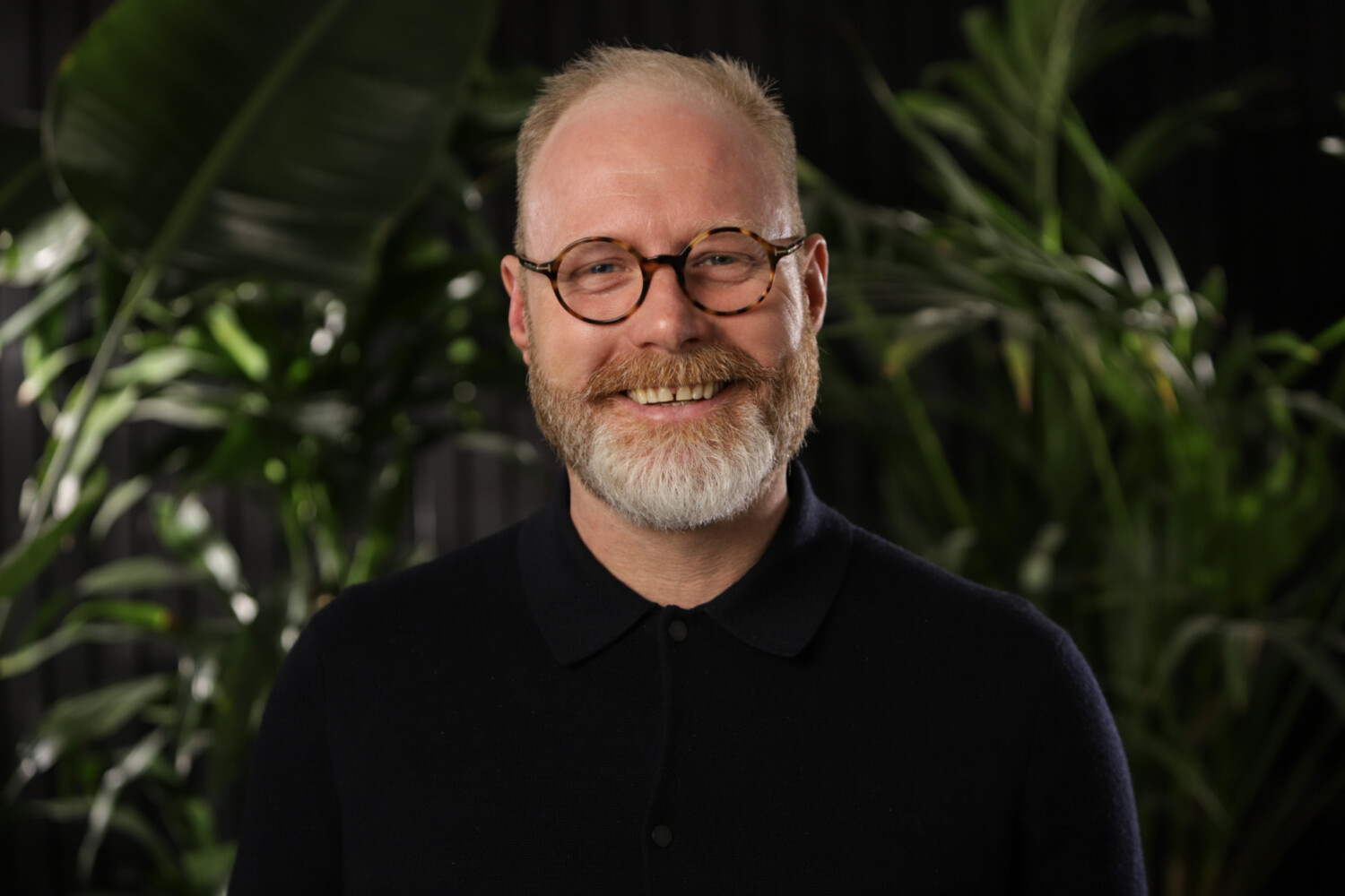 Middle-aged man with grey beard and glasses wearing black shirt, smiling at camera with green plants in background.