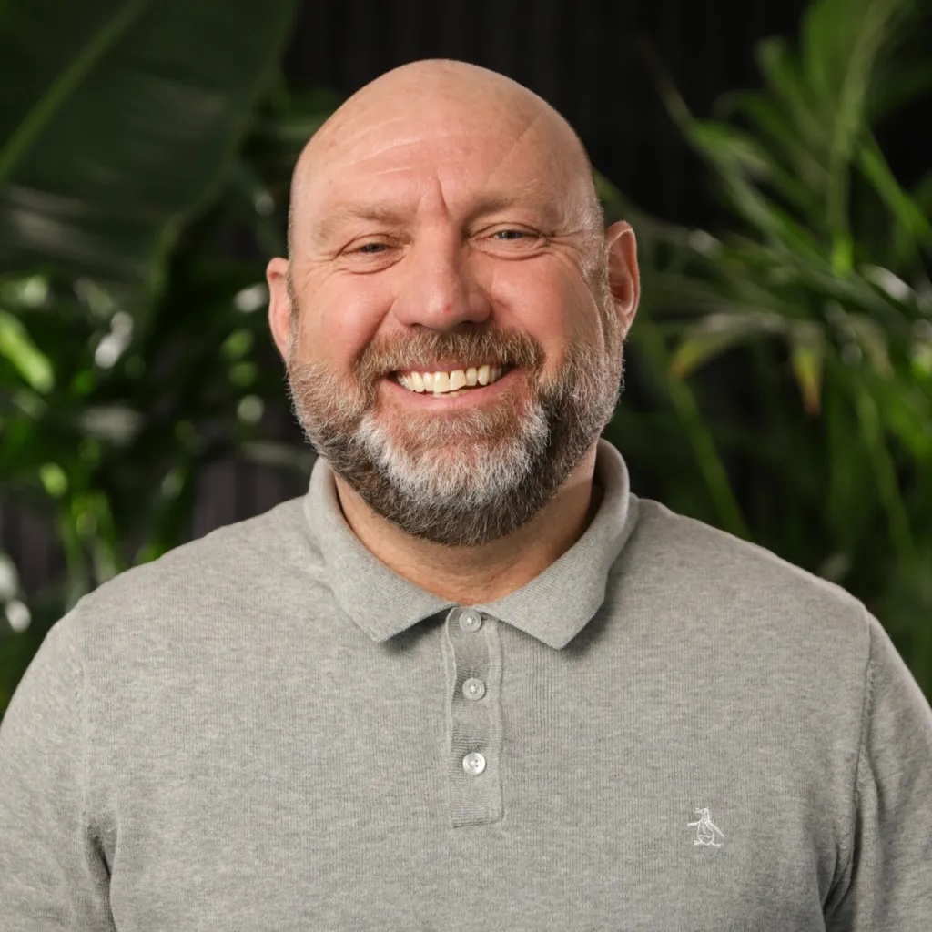 Bald man with grey beard smiling, wearing light grey polo shirt with small logo, green foliage background.